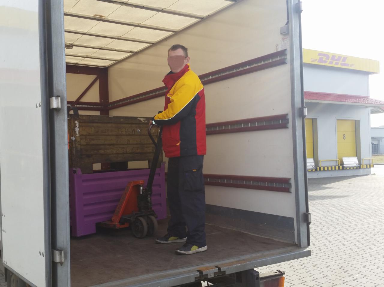 Pracownik w kurtce DHL manewruje wózkiem paletowym z ładunkiem wewnątrz ciężarówki. W tle budynek DHL z żółtymi drzwiami i logo.
