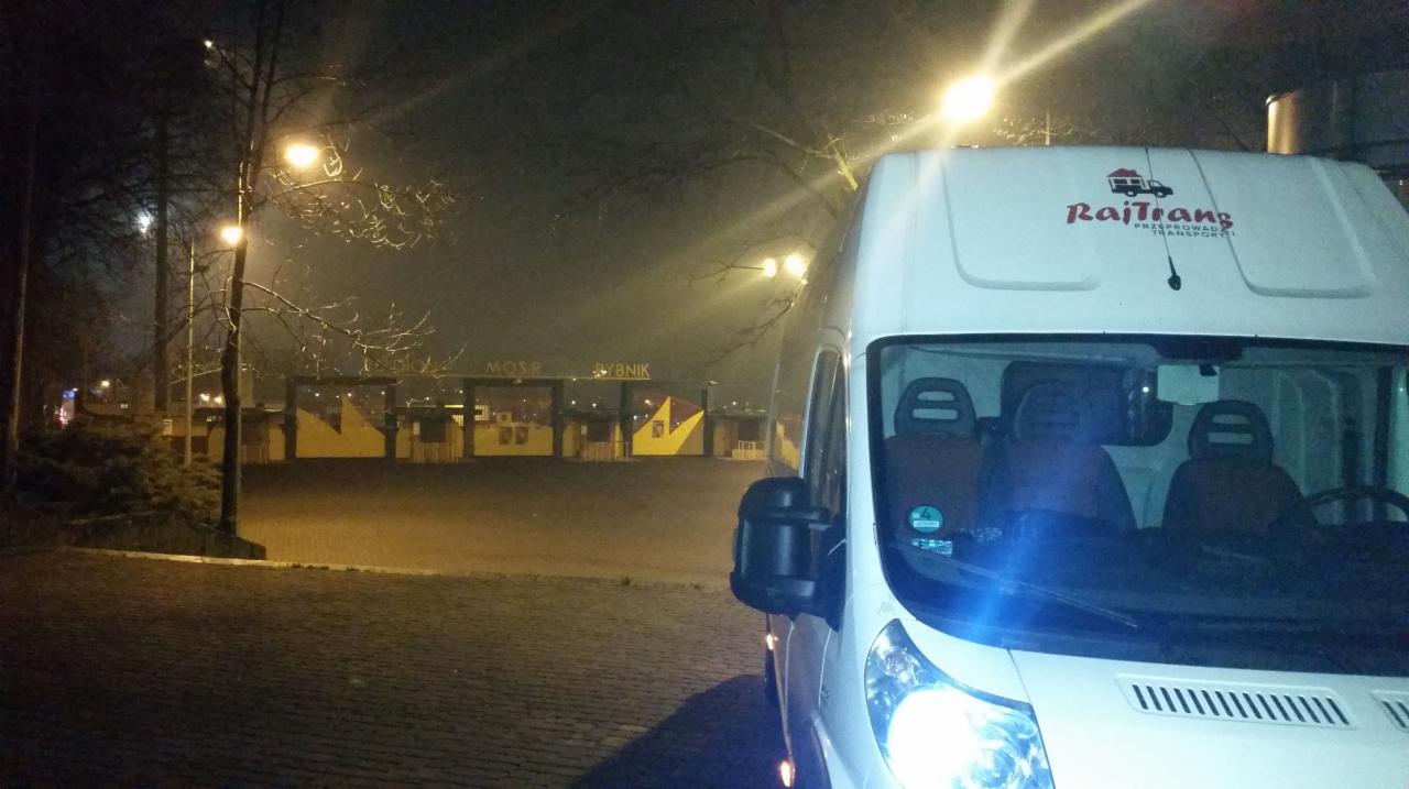 Biały bus firmy RajTrans z logo, zaparkowany nocą na brukowanej nawierzchni, w tle oświetlony budynek z napisem MOSiR Rybnik. Widoczne drzewa bez liści.