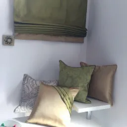 Dekoracyjne poduszki w odcieniach beżu i zieleni, ułożone na białym stoliku obok zielonej rolety rzymskiej z beżowym pasem na dole, zamontowanej na ścianie.