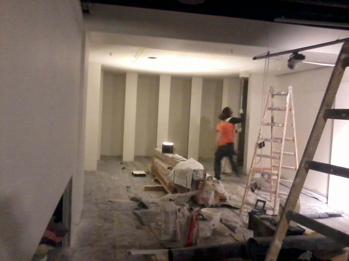 Wnętrze w trakcie remontu: pracownik w pomarańczowej koszulce maluje ścianę z pionowymi pasami. Widoczne narzędzia, drabiny i materiały budowlane.