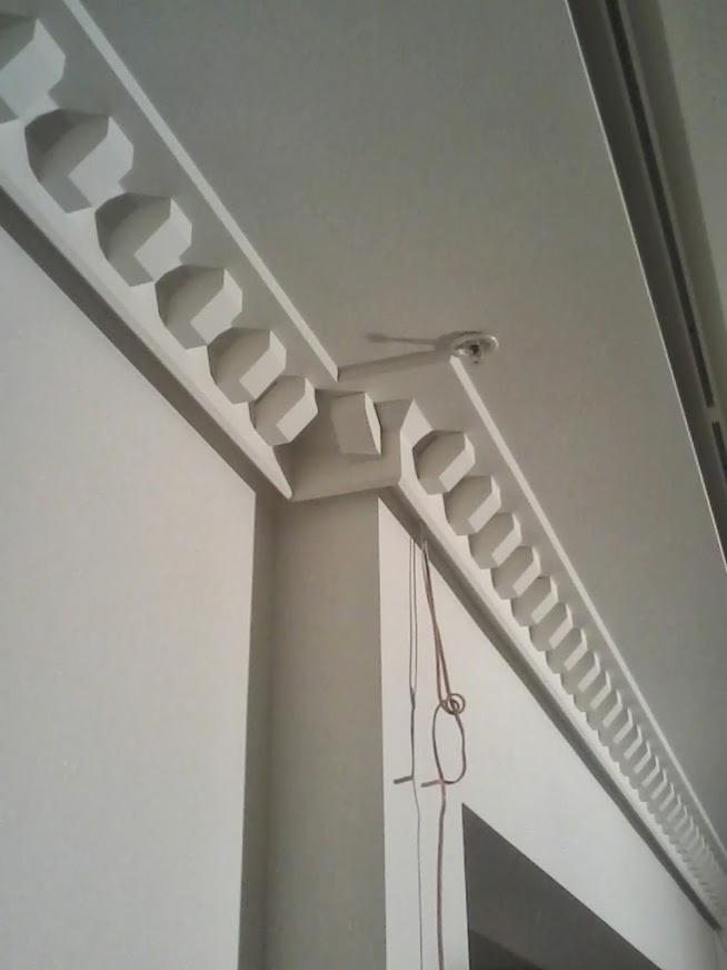 Detal wykończenia wnętrza: białe, ozdobne listwy sufitowe z geometrycznym wzorem, zamontowane na styku ściany i sufitu. Widoczna lampa sufitowa i fragment okna.