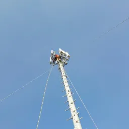 Pracownik w pomarańczowym kombinezonie na szczycie wieży telekomunikacyjnej, montujący lub serwisujący anteny sektorowe, widok z dołu na tle błękitnego nieba.