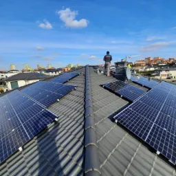 Instalacja paneli słonecznych na dachu domu jednorodzinnego z widocznym pracownikiem w odzieży roboczej i anteną satelitarną na kominie, w tle zabudowa miejska i błękitne niebo.