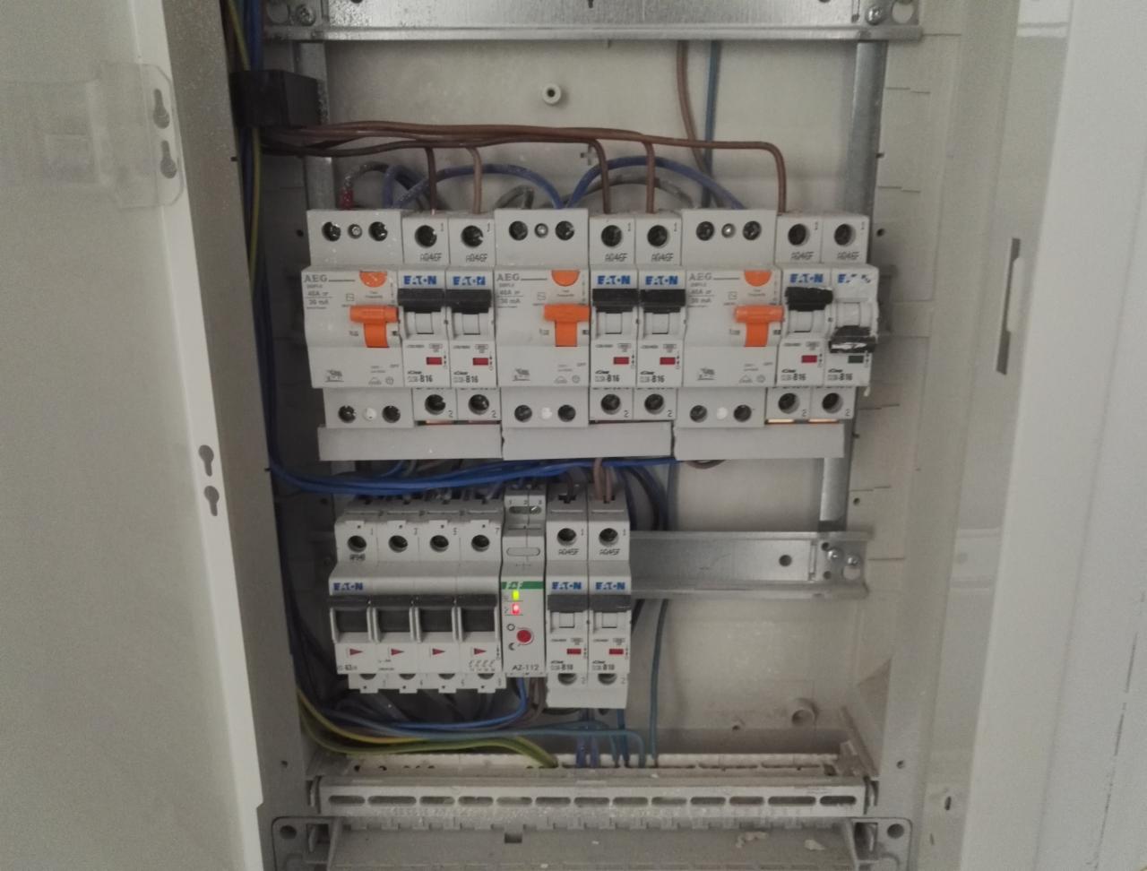Otwarta skrzynka elektryczna z widocznymi bezpiecznikami AEG i Eaton, różnokolorowe przewody podłączone do listwy zaciskowej, jeden bezpiecznik z czerwoną diodą LED sygnalizującą stan.