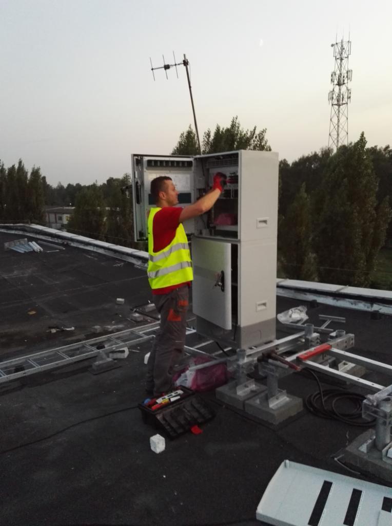 Elektryk w kamizelce odblaskowej, na dachu, pracuje przy otwartej szafie elektrycznej, widoczna antena i wieża telekomunikacyjna w tle.