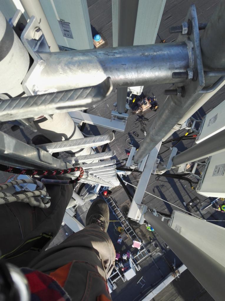 Widok z góry na metalową konstrukcję na dachu, z perspektywy osoby stojącej na drabinie, widoczne nogi w roboczych spodniach i butach, liny zabezpieczające oraz ekipa pracująca na dachu budynku.