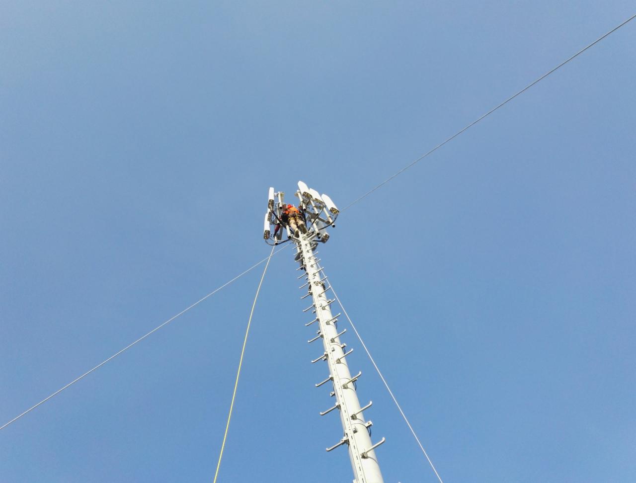 Pracownik w pomarańczowym kombinezonie na szczycie wieży telekomunikacyjnej, montujący lub serwisujący anteny sektorowe, widok z dołu na tle błękitnego nieba.