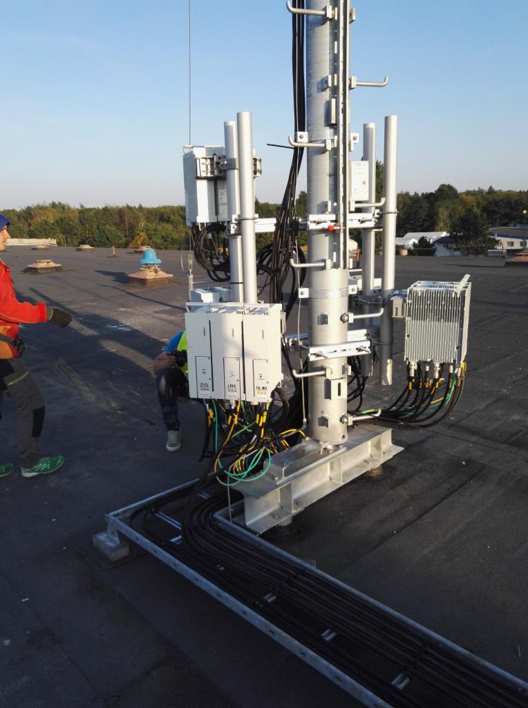 Instalacja nowej stacji bazowej telefonii komórkowej na płaskim dachu budynku, widoczne moduły, kable i pracownicy w trakcie montażu.