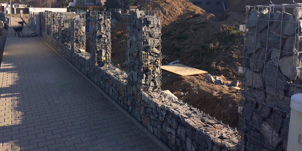 Gabiony z kamienia, tworzące ogrodzenie wzdłuż chodnika z kostki brukowej. Widoczny etap budowy z ziemią w tle i fragmentami materiałów budowlanych.