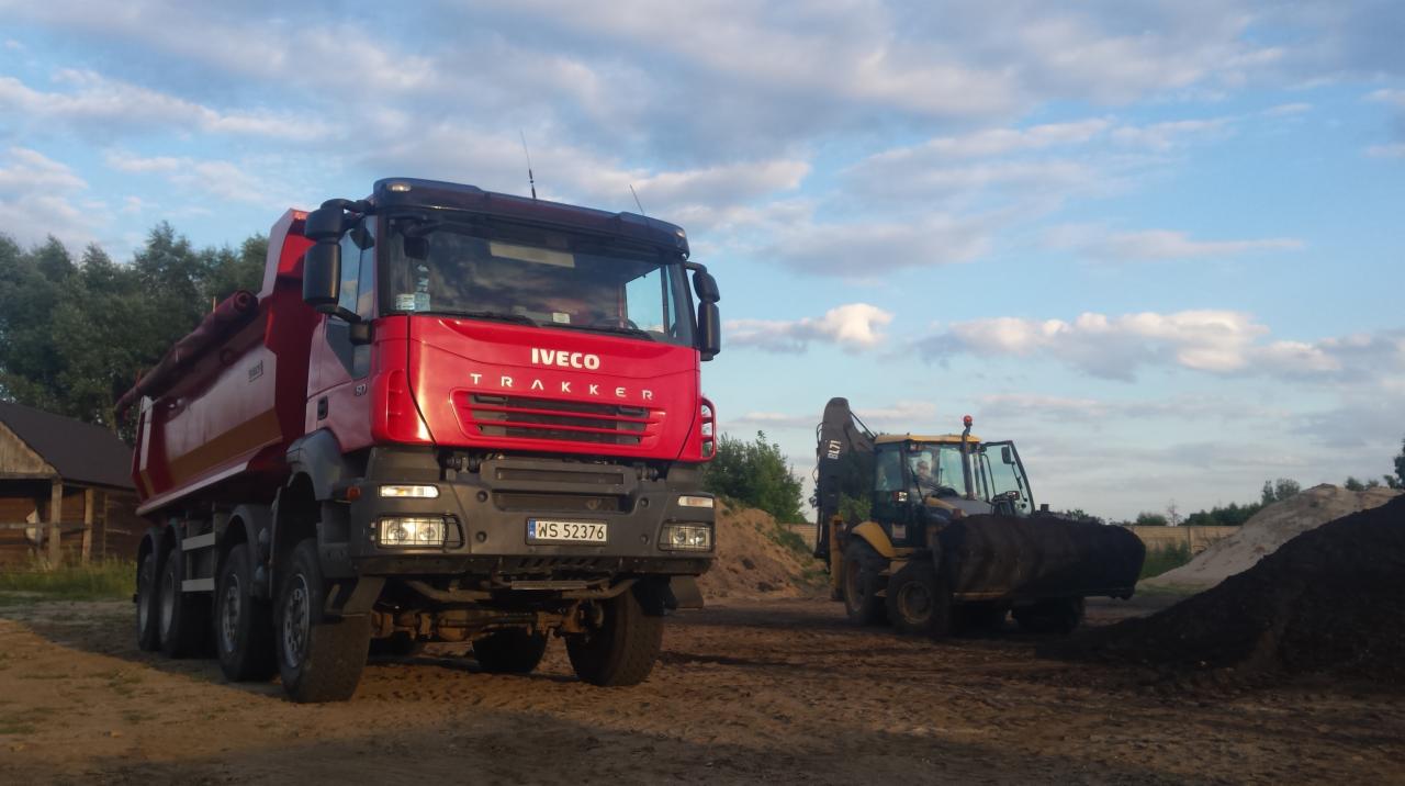 Czerwona wywrotka Iveco Trakker z numerem WS 52376 i żółta koparko-ładowarka na placu budowy pod błękitnym niebem z obłokami.