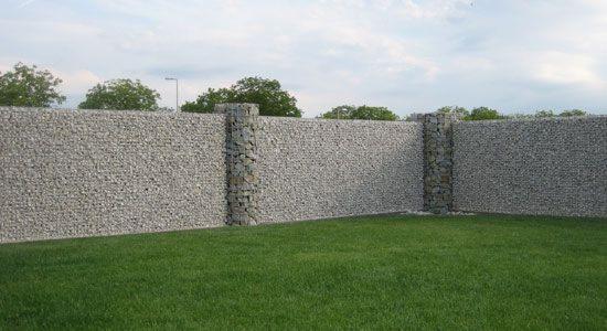 Gabionowy mur z kamiennymi filarami, otoczony zielonym trawnikiem w słoneczny dzień. Konstrukcja ogrodzenia z koszy wypełnionych kamieniami, widok z poziomu trawy.