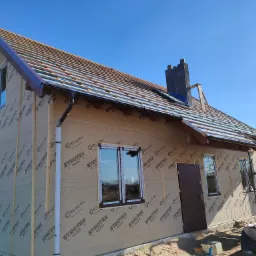 Dom szkieletowy w trakcie budowy, widok z zewnątrz na ocieploną ścianę z folią Strotex, zamontowane okna z brązowymi ramami, przygotowane podłoże pod dach z widocznym kominem.