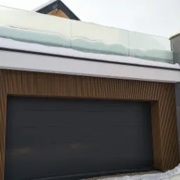 Nowoczesny garaż z szarą bramą segmentową, okładziną z drewnianych listew i szklaną balustradą na dachu, częściowo pokrytą śniegiem.