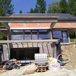 KBKG Budownictwo Grzegorz Luranc - Surowy stan domu w trakcie budowy z widocznymi bloczkami, otworami okiennymi i betonową konstrukcją szkieletową, na tle zieleni i błękitnego nieba.