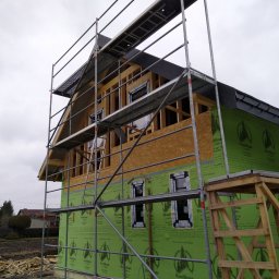 KBKG Budownictwo Grzegorz Luranc - Dom w trakcie budowy z widoczną konstrukcją szkieletową, ocieplony zieloną membraną, otoczony rusztowaniami i stertą drewna na ziemi.