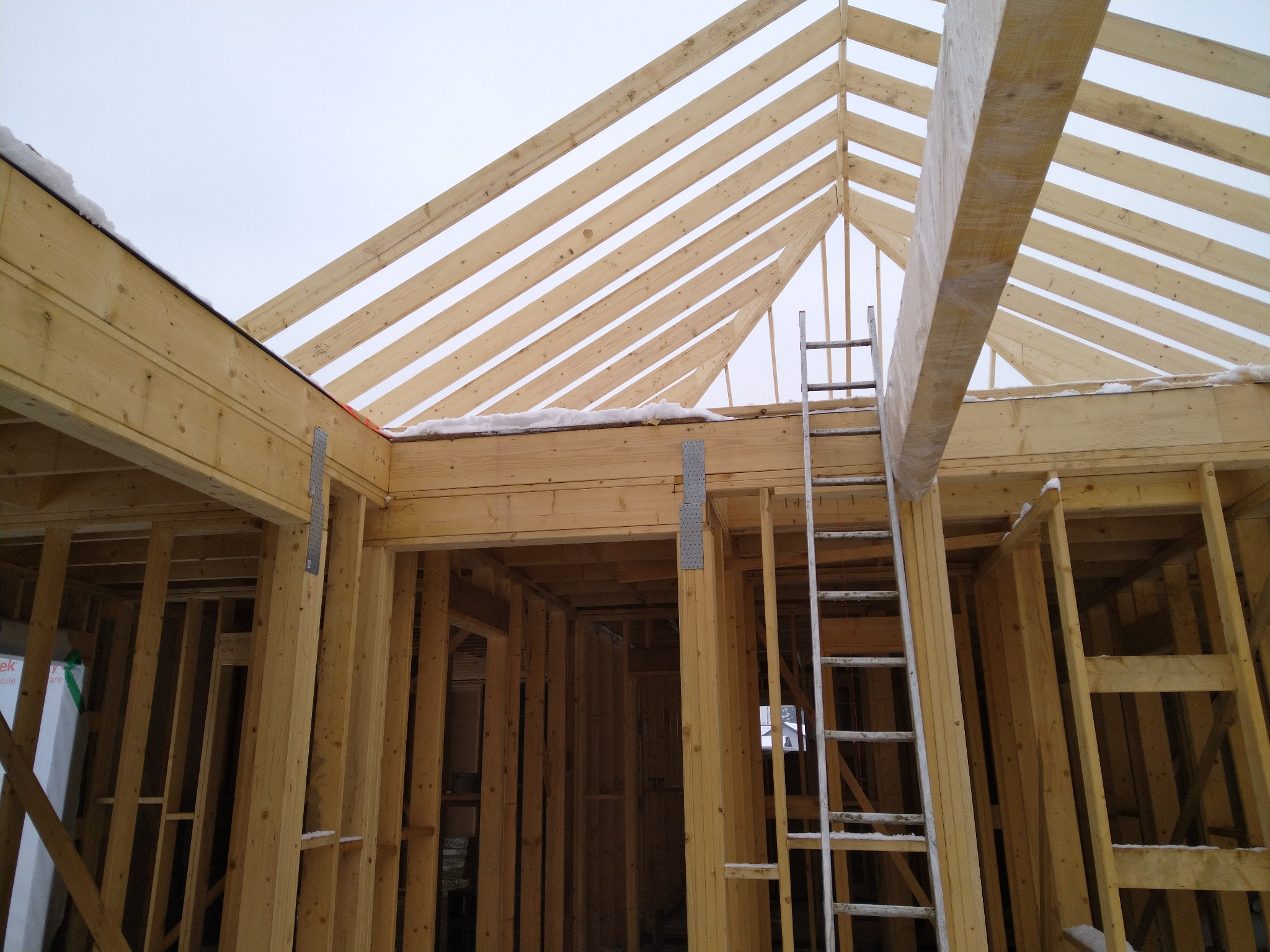 Surowa konstrukcja drewniana domu w trakcie budowy, widoczne krokwie dachu, ściany szkieletowe i drabina oparta o konstrukcję, lekki śnieg na elementach dachu.