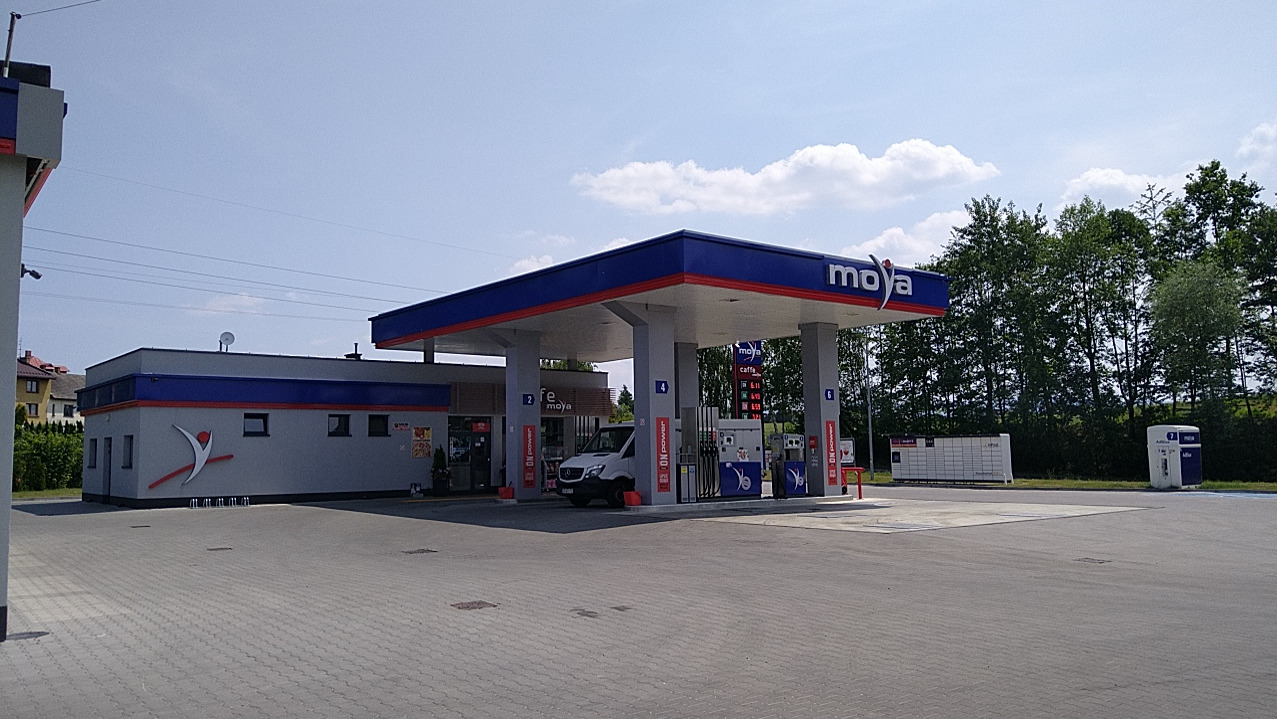 Stacja paliw Moya z zaparkowanym białym vanem, widok na budynek stacji, dystrybutory i automat paczkowy na placu z kostki brukowej.