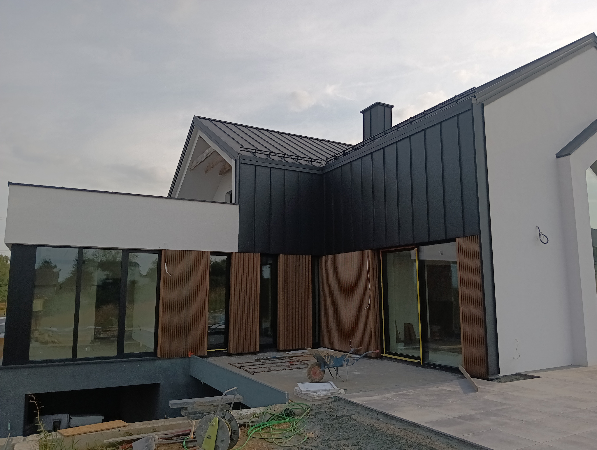 Nowoczesny dom z ciemną blachą na dachu i drewnianymi panelami elewacyjnymi, widok z zewnątrz na taras w trakcie wykańczania, z widocznymi narzędziami budowlanymi i taczką.