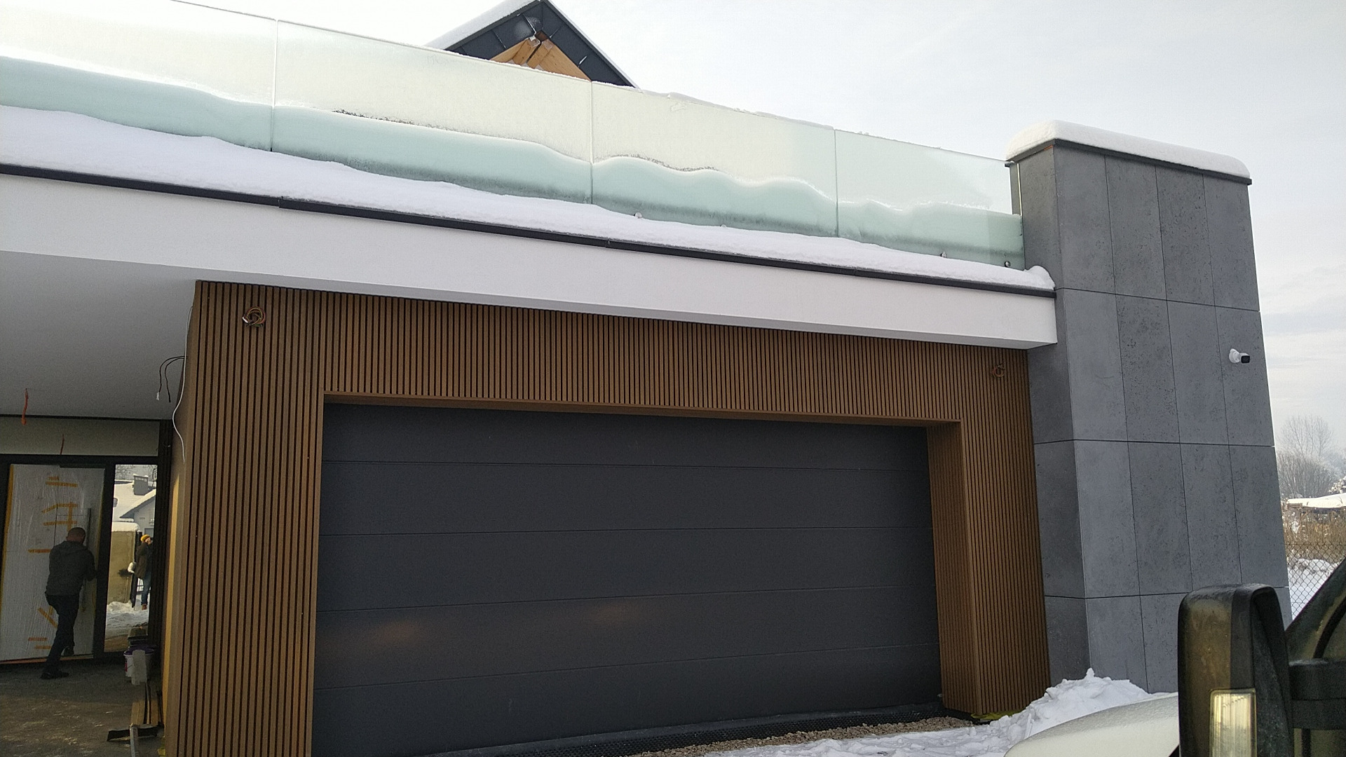 Nowoczesny garaż z szarą bramą segmentową, okładziną z drewnianych listew i szklaną balustradą na dachu, częściowo pokrytą śniegiem.