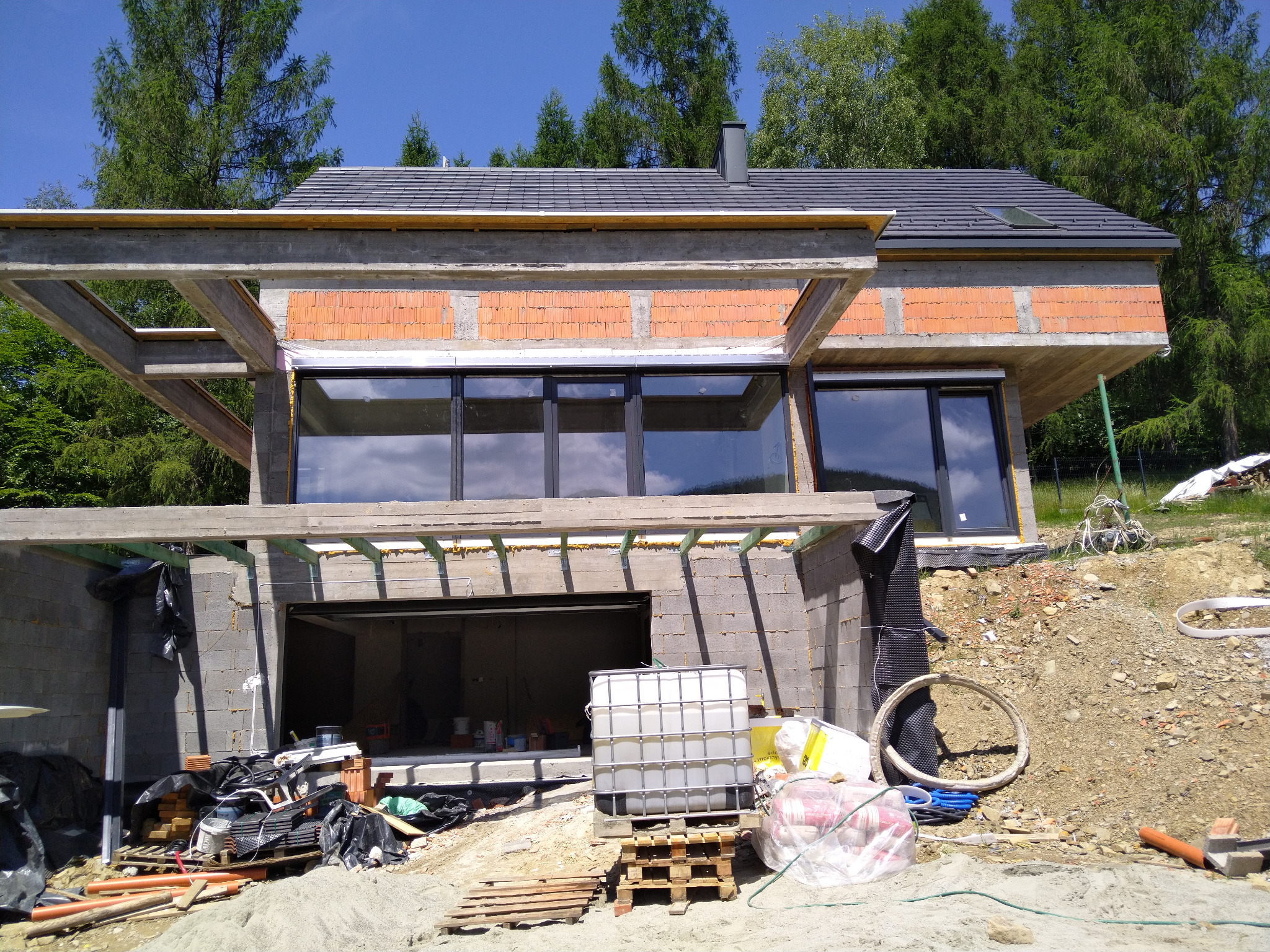 Surowy stan domu w trakcie budowy z widocznymi bloczkami, otworami okiennymi i betonową konstrukcją szkieletową, na tle zieleni i błękitnego nieba.