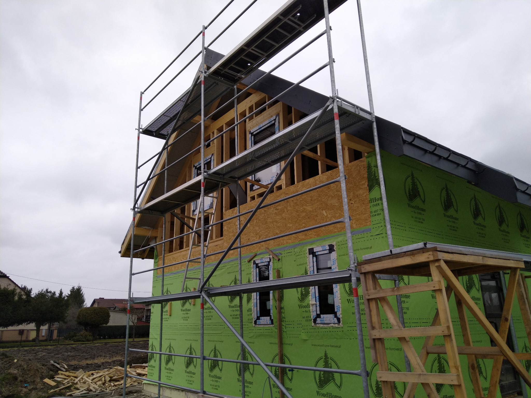 Dom w trakcie budowy z widoczną konstrukcją szkieletową, ocieplony zieloną membraną, otoczony rusztowaniami i stertą drewna na ziemi.