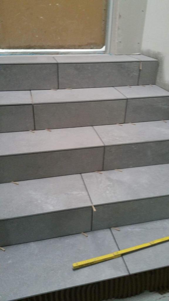 Wykończone schody z szarych płytek, z widocznymi krzyżykami dystansowymi i żółtą miarką na dolnym stopniu. Ujęcie z dołu, widok na trzy stopnie.