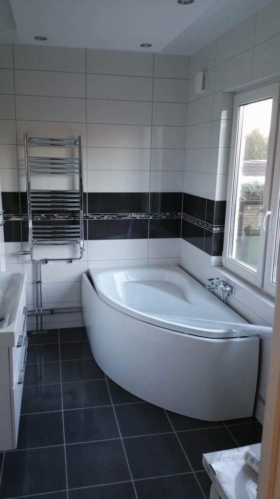 Nowoczesna łazienka z asymetryczną wanną narożną, grzejnikiem drabinkowym i czarno-białymi płytkami. Minimalistyczny design, ciemna podłoga, jasne ściany.