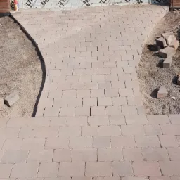 Ułożona ścieżka z kostki brukowej prowadząca do wejścia budynku. Widoczne niedokończone obrzeża i zapas kostki. Na ścianie budynku widoczna folia izolacyjna.