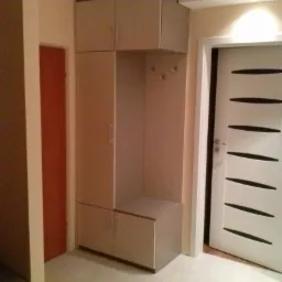 Jasna szafa wnękowa z siedziskiem i haczykami na ubrania, dopasowana do wnętrza przedpokoju, obok białe drzwi z poziomymi, czarnymi paskami.