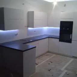 Nowoczesna, biała kuchnia z ciemnym blatem i subtelnym oświetleniem LED. Uwagę przyciąga minimalistyczny design i brak uchwytów w szafkach.