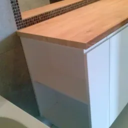 Biała szafka łazienkowa z blatem z jasnego drewna i otwartą półką, widoczna mozaika na ścianie, wnętrze łazienki.