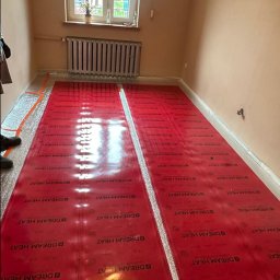 USŁUGI ELEKTRO-INSTALACYJNE SEBASTIAN WAWRZYNEK - Montaż folii grzewczej Dream Heat na podłodze w pomieszczeniu w Bierawie, widoczny grzejnik pod oknem i fragment instalatora w lewym dolnym rogu.