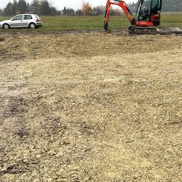 USŁUGI ELEKTRO-INSTALACYJNE SEBASTIAN WAWRZYNEK - Przygotowanie terenu pod budowę: wyrównany teren z użyciem mini koparki Kubota i ubijarki, widoczne samochody w tle.