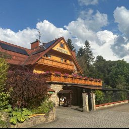 USŁUGI ELEKTRO-INSTALACYJNE SEBASTIAN WAWRZYNEK - Drewniany pensjonat z czerwonym dachem i panelami słonecznymi, balkonem ozdobionym kwiatami i kamienną elewacją, otoczony zielenią i brukowanym podjazdem.