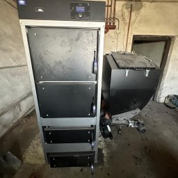 USŁUGI ELEKTRO-INSTALACYJNE SEBASTIAN WAWRZYNEK - Nowoczesny piec CO z podajnikiem, widoczne rury instalacji grzewczej na tle surowej ściany w kotłowni