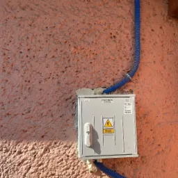 Szara skrzynka elektryczna z ostrzeżeniem o wysokim napięciu, zamontowana na nierównej, pomarańczowej ścianie w Bierawie, z niebieskimi rurami ochronnymi doprowadzającymi przewody.