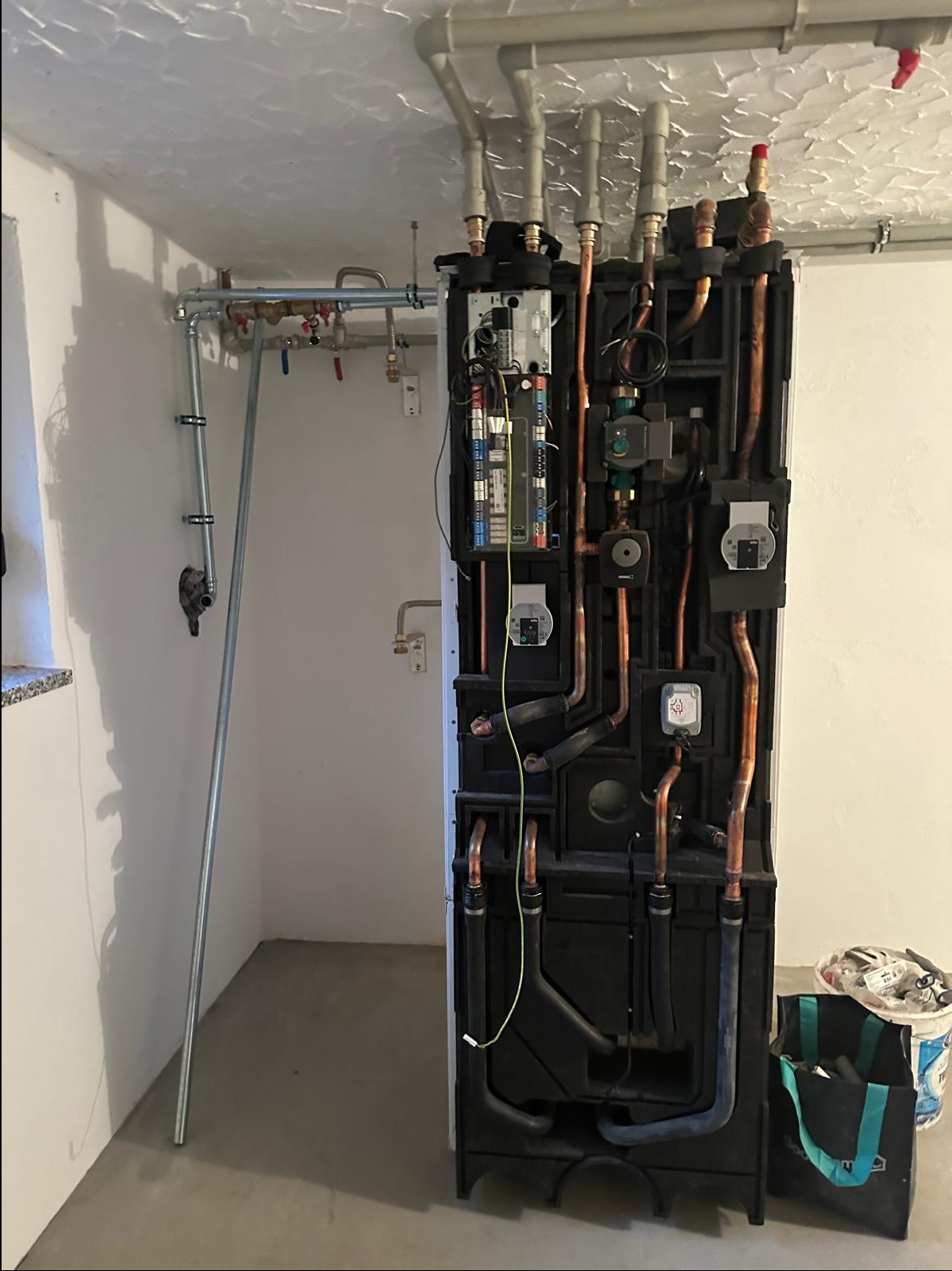 Nowoczesny, kompaktowy piec grzewczy z widocznymi miedzianymi rurami i licznymi podłączeniami, zainstalowany w pomieszczeniu technicznym z szarymi rurami pod sufitem i torbą narzędziową obok.