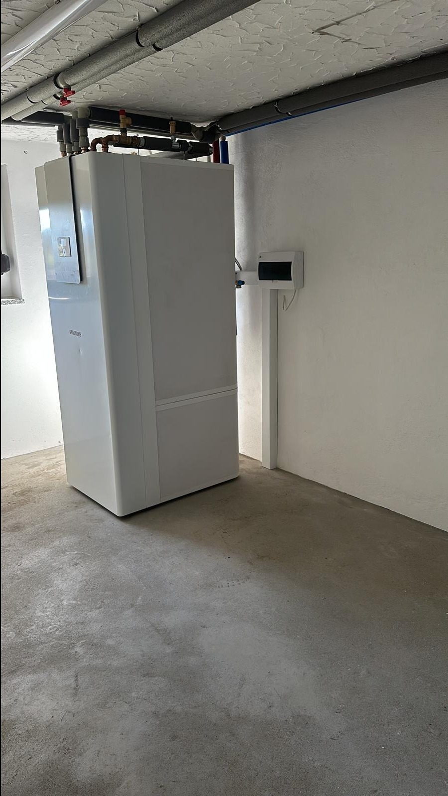 Biała pompa ciepła w piwnicy, widoczne rury izolowane na suficie i skrzynka elektryczna na ścianie.
