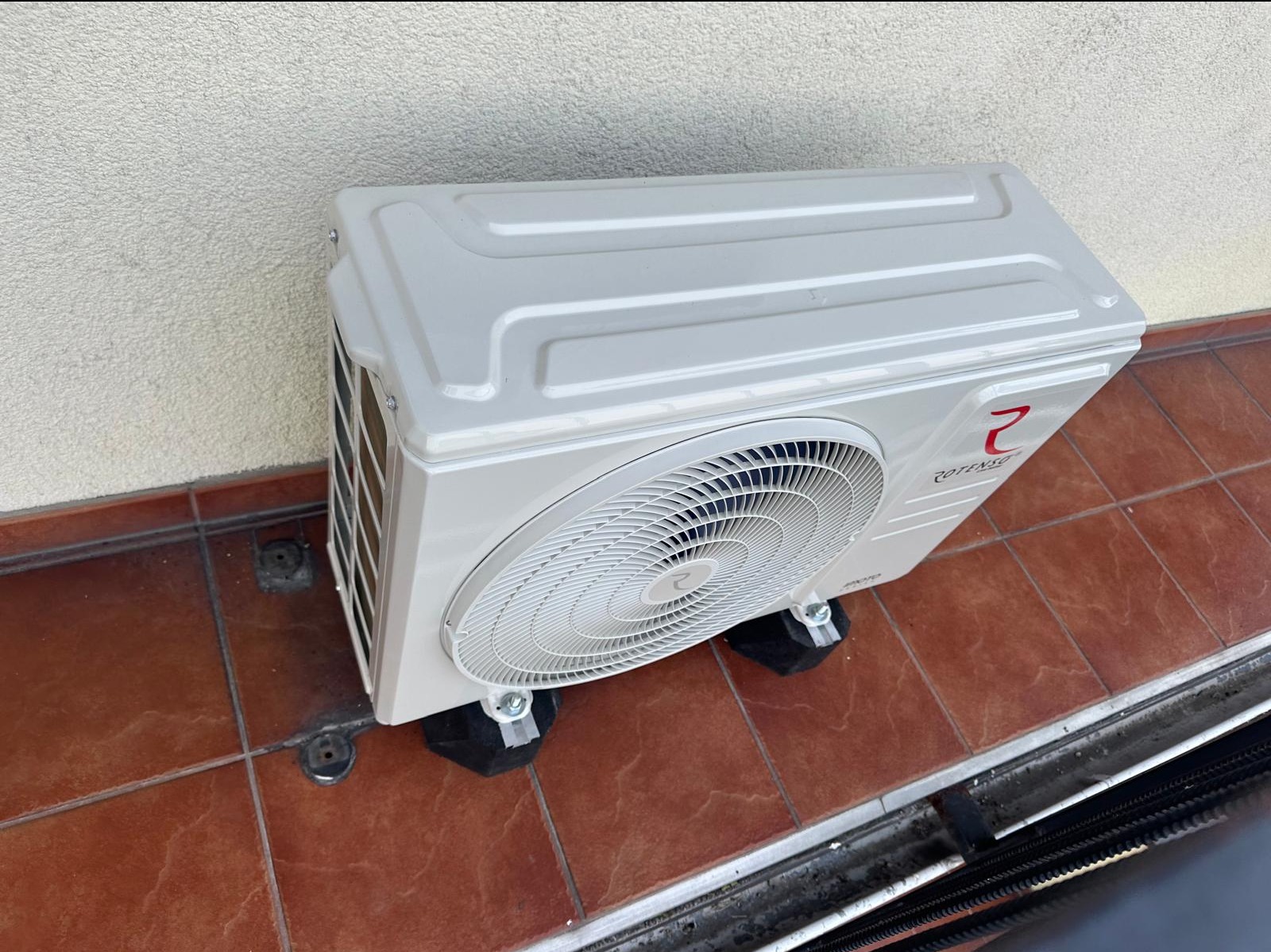 Zewnętrzna jednostka klimatyzatora marki Rotenso, umieszczona na czarnych podkładkach na brązowych płytkach tarasu, widoczna ściana budynku z jasnym tynkiem.