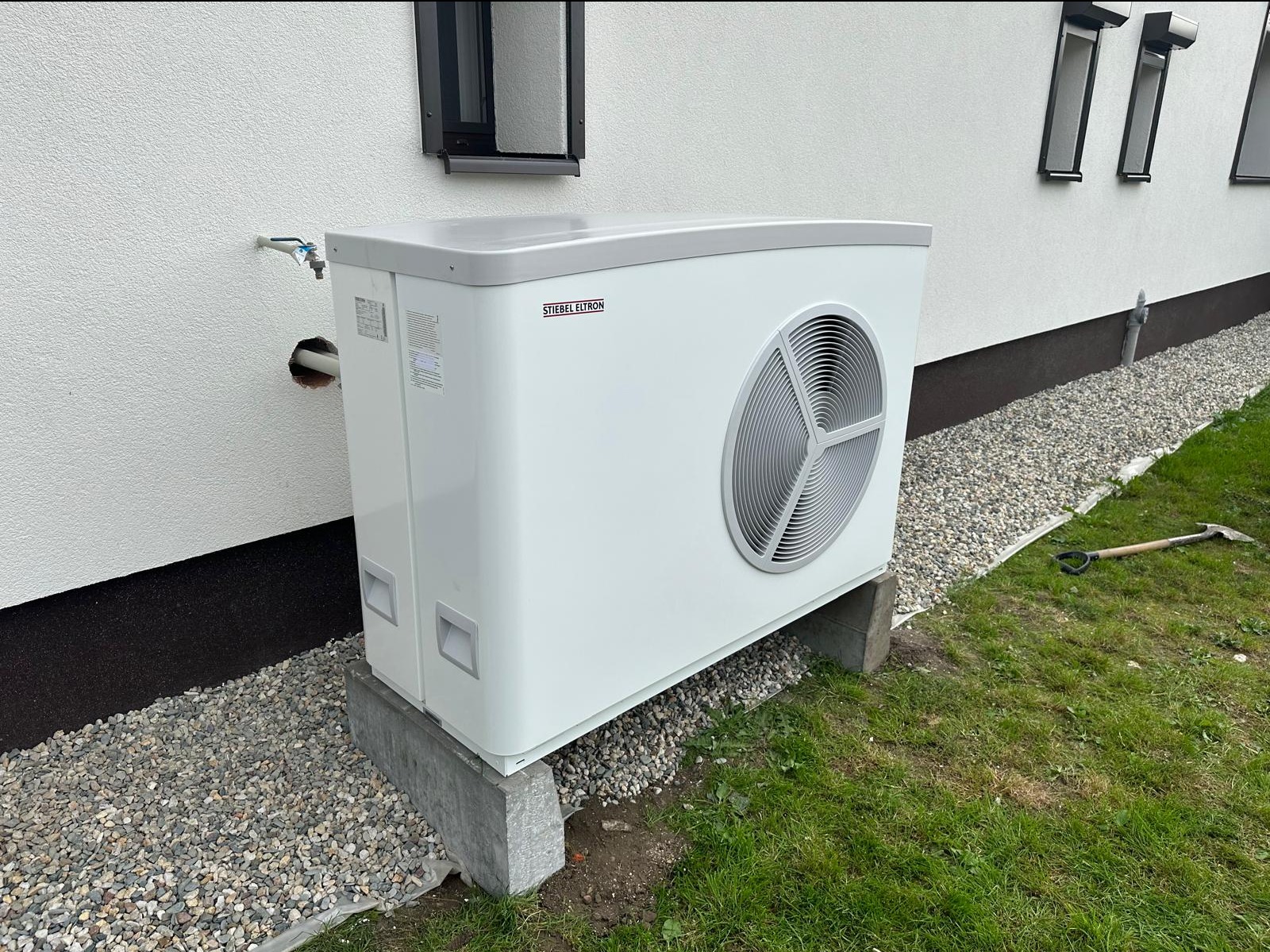 Biała pompa ciepła marki Stiebel Eltron, umieszczona na betonowych fundamentach przy ścianie domu z oknami, obok widoczny kran na ścianie i rura wchodząca do budynku, w tle trawnik i kamienie.