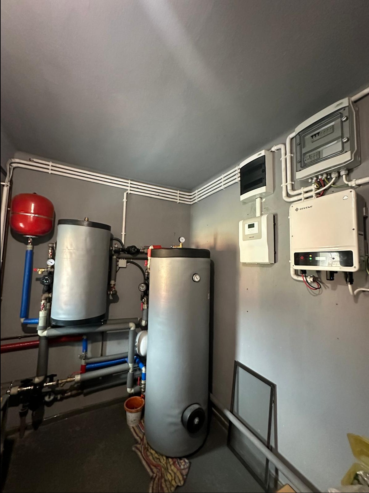 W kotłowni dwa szare zbiorniki akumulacyjne z czerwonym naczyniem wzbiorczym, instalacja rurowa z zaworami, na ścianie rozdzielnica elektryczna i inwerter solarny, na podłodze wiadro i szmata.