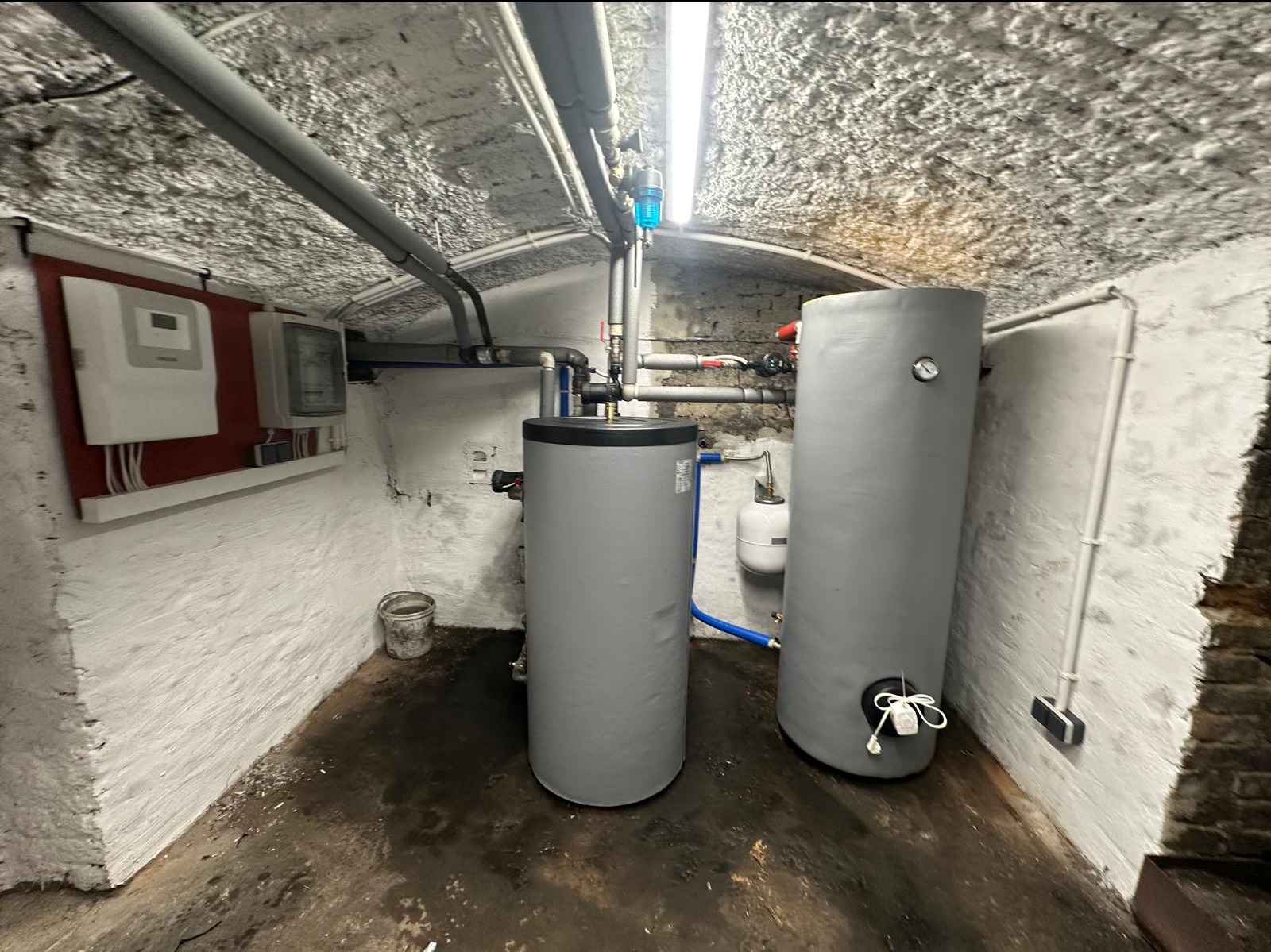 Dwa szare, cylindryczne zasobniki ciepłej wody w piwnicy o nierównych ścianach i suficie, z widocznymi rurami i instalacją hydrauliczną, oraz białą skrzynką sterującą na ścianie.