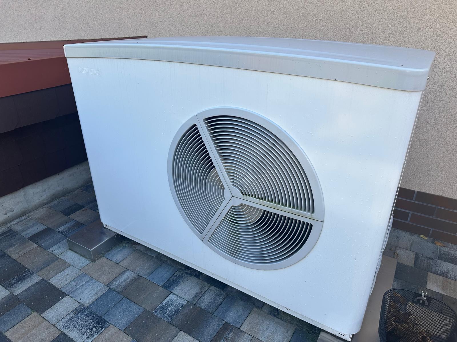Zewnętrzna jednostka pompy ciepła powietrze-woda, umieszczona na kostce brukowej, z widocznym odpływem kondensatu i fragmentem rynny w tle.