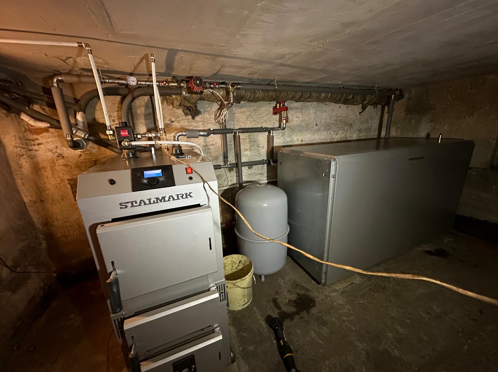 Kotłownia w piwnicy z piecem marki Stalmark, zbiornikiem wyrównawczym i zasobnikiem ciepłej wody użytkowej, widoczne rury i instalacje.