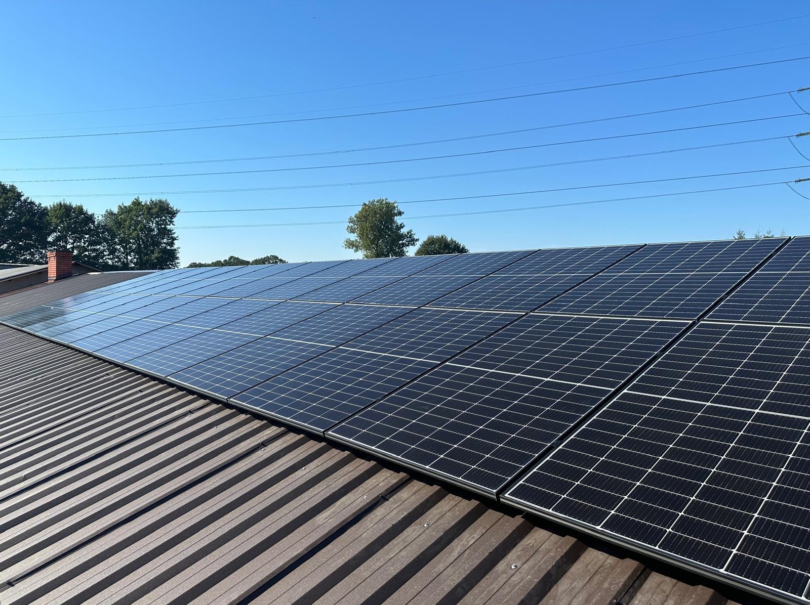 Panele słoneczne na metalowym dachu domu jednorodzinnego, widoczne linie wysokiego napięcia i błękitne niebo.