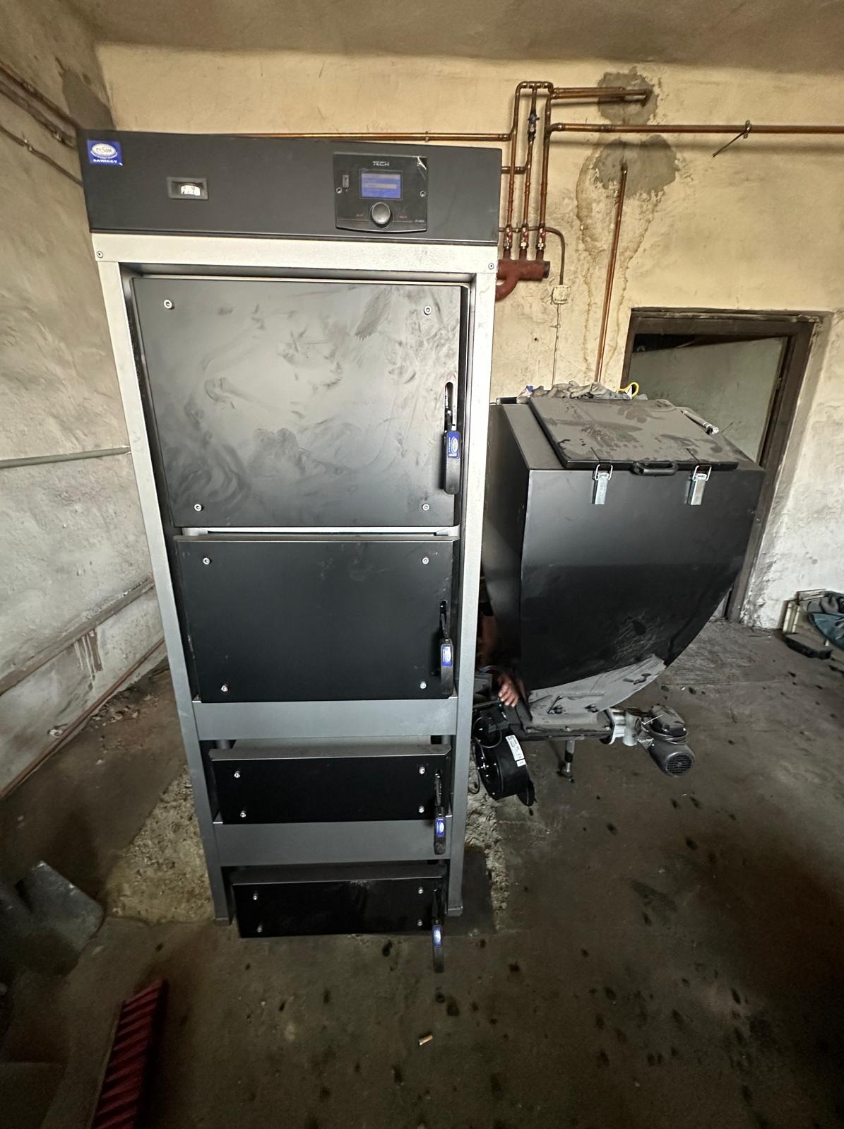 Nowoczesny piec CO z podajnikiem, widoczne rury instalacji grzewczej na tle surowej ściany w kotłowni