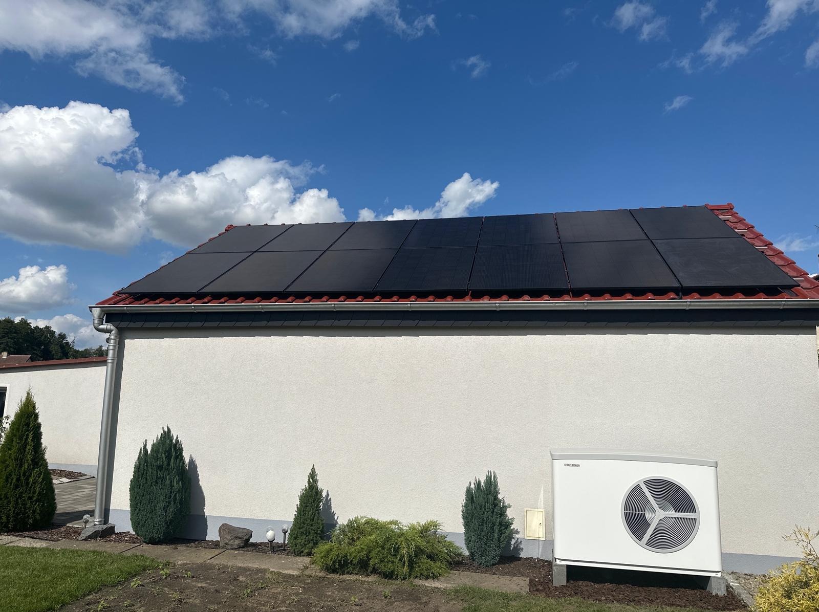Czarny panel fotowoltaiczny na czerwonym dachu domu jednorodzinnego, obok biała pompa ciepła, błękitne niebo z chmurami w tle.