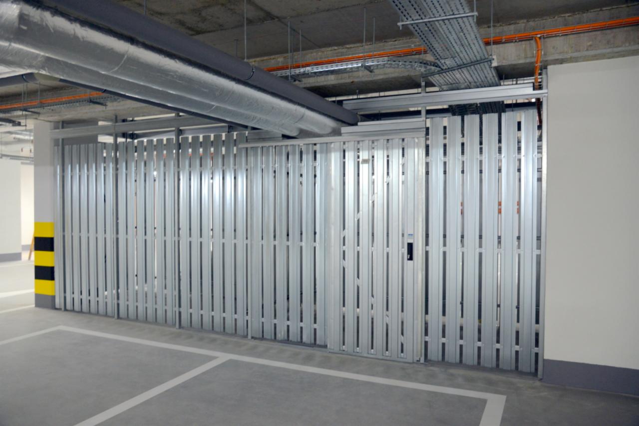 Metalowa, przesuwna brama garażowa w parkingu podziemnym, z widocznymi instalacjami pod sufitem i oznaczeniami na słupie. Zamknięta konstrukcja z wąskimi panelami.