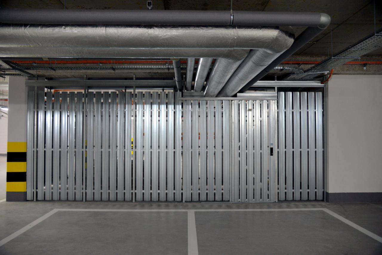 Metalowa, składana brama garażowa w hali garażowej, widoczne rury wentylacyjne pod sufitem i oznaczenia na filarze. Minimalistyczny design, szara kolorystyka.