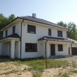 Dwukondygnacyjny dom z jasną elewacją, ciemną dachówką i brązowymi roletami zewnętrznymi, widok z zewnątrz na tle zieleni.